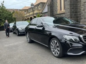 A black Mercedes-Benz E-Class limousine lead car in a procession with a chauffeur.